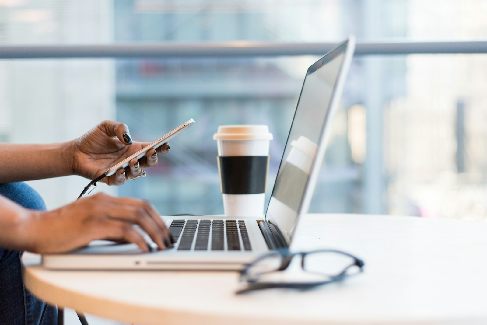 Person using smartphone and laptop on table to set up smart home ecosystem with coffee cup