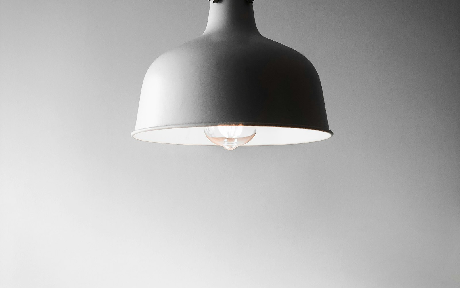 A gray metal pendant lamp with a clear glass bulb glowing against a plain wall.